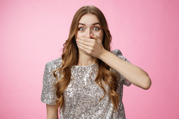 A girl against a pink background with her hand covering her mouth area and lip filler swelling, looking shocked